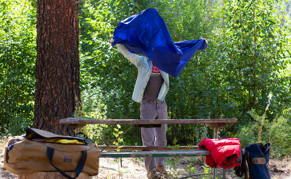 A Large Shepherd's Wool Utility Cloth in Neela Blue being shaken into the air to spread as a table cloth on a picnic table at a campsite.